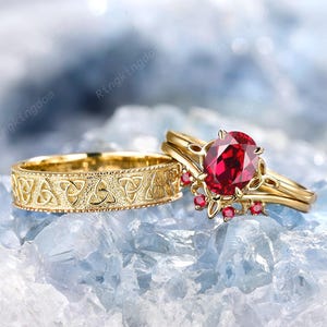 May include: Two gold rings. One ring has a Celtic knot pattern. The other ring features a large oval red gemstone and smaller red gemstones. The rings are set against a light blue background.