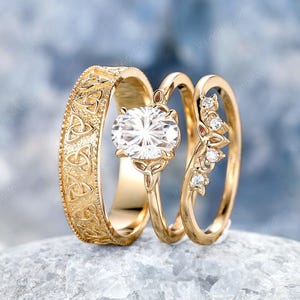 Könnte beinhalten: Drei goldene Eheringe. Der Verlobungsring hat einen großen, oval geschliffenen Diamanten. Der Ehering hat ein kompliziertes keltisches Knotendesign. Der dritte Ring hat kleine Diamanten. Alle Ringe liegen auf einer hellblauen Oberfläche.
