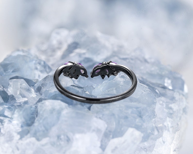 Pode incluir: Um anel preto com duas pedras preciosas roxas engastadas em um design de folhas. O anel est&aacute; apoiado em um leito de cristais brancos.