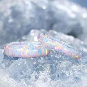 Puede incluir: Dos anillos de ópalo blanco con destellos iridiscentes. Uno es una banda ancha y el otro una banda más fina. Los anillos se exhiben sobre un lecho de cristales azul claro. Los anillos tienen un acabado liso y pulido.