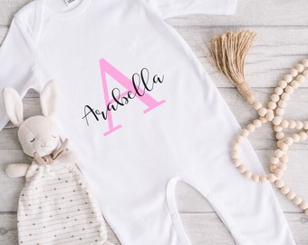 May include: White baby onesie with a pink letter "A" and the name "Arabella" in black script. The onesie is laid flat on a light wood surface, next to a stuffed bunny and wooden beads.