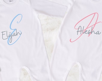 May include: Two white baby onesies with long sleeves, each personalized with a name. One features "Elijah" in blue script with a blue "S", the other "Alesha" in pink script with a pink "A". Wooden baby toys are on either side.