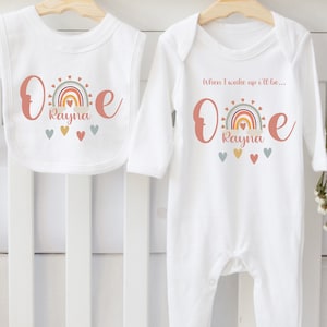 Puede incluir: Conjunto de babero y body para bebé blancos con un arcoíris y el nombre "Rayna". El babero y el body tienen la palabra "One" en color melocotón. El body tiene el texto "When I wake up I'll be..."