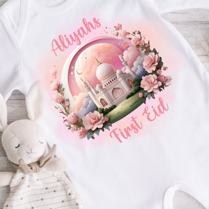 May include: White baby onesie with a pink and white illustration of a mosque, moon, and flowers. The text "Aliyahs First Eid" is written in pink script. A plush bunny toy is in the lower left corner.