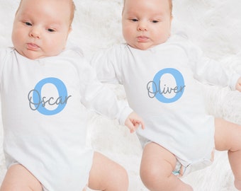 May include: Two white baby onesies with long sleeves. One has the name "Oscar" and a blue "O" on the front. The other has the name "Oliver" and a blue "O". The babies are lying on a white, fluffy surface.
