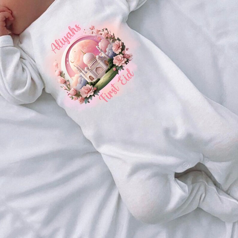 May include: White baby sleeper with long sleeves. The sleeper features a pink and white graphic of a mosque, flowers, and the text "Aliyahs First Eid". The design is centered on the chest.