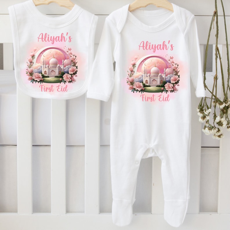 May include: White baby bib and onesie set. Both items feature a pink and white graphic of a mosque, flowers, and the text "Aliyah's First Eid". The onesie has long sleeves and footies.