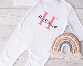 May include: White baby onesie with long sleeves, featuring a large pink letter "H" and the name "Halie" in cursive script. A small heart is drawn next to the name. A decorative rainbow in neutral colors sits beside the onesie.