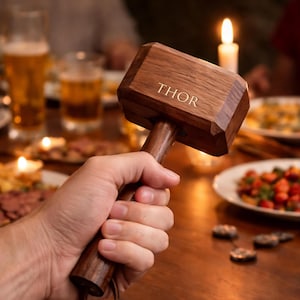 May include: A wooden hammer with the word "THOR" engraved on the head. The hammer is held in a person's hand, with a dining table set with food and drinks in the background. The hammer is a dark brown color.