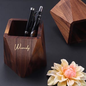 May include: A wooden pen holder with a geometric design, holding two black pens. The holder has the word "Weorndy" in gold lettering. A second, similar wooden piece is nearby, along with a light pink and yellow flower.