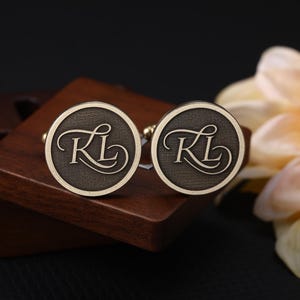 May include: A pair of round cufflinks with a dark bronze finish, each featuring the initials "KL" in an elegant script. The cufflinks are set against a dark wooden box and a blurred flower.