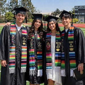 May include: Four graduates in black gowns and mortarboards, each with a colourful stole. The stoles display "Class of 2026" and feature rainbow stripes and floral embroidery. They are standing on a green field, with a sports stadium in the background.