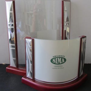 Pode incluir: Duas molduras decorativas para fotos. As molduras têm uma frente de vidro transparente curvada e uma base de madeira vermelha escura. As laterais são acentuadas com metal prateado. O logotipo "ARGENTI SIMA MADE IN ITALY" é visível na frente de uma moldura.