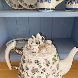 May include: A white ceramic teapot featuring a colourful floral pattern with pink, green, and purple flowers. The lid is designed as a miniature table set with a tiny teapot, creamer, cups, and plates, creating a whimsical tea party scene.