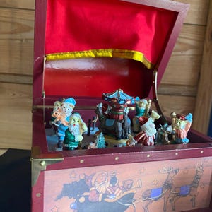 Puede incluir: Una caja de música de madera roja con tapa abatible, que presenta una escena navideña. En el interior, se ven figuras en miniatura de Papá Noel, elfos y un carrusel. El exterior de la caja está decorado con una escena de Papá Noel y trineo de renos.