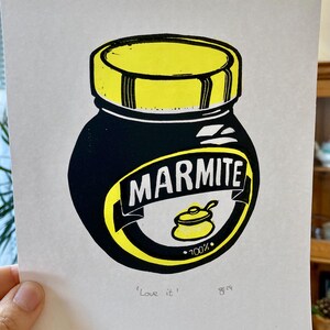 May include: A black and yellow linocut print of a Marmite jar. The jar has a yellow lid and the text "Marmite" in white letters. The text "100%" is printed in black below a yellow pot with a spoon. The text "'Love it'" is printed in black at the bottom of the print.