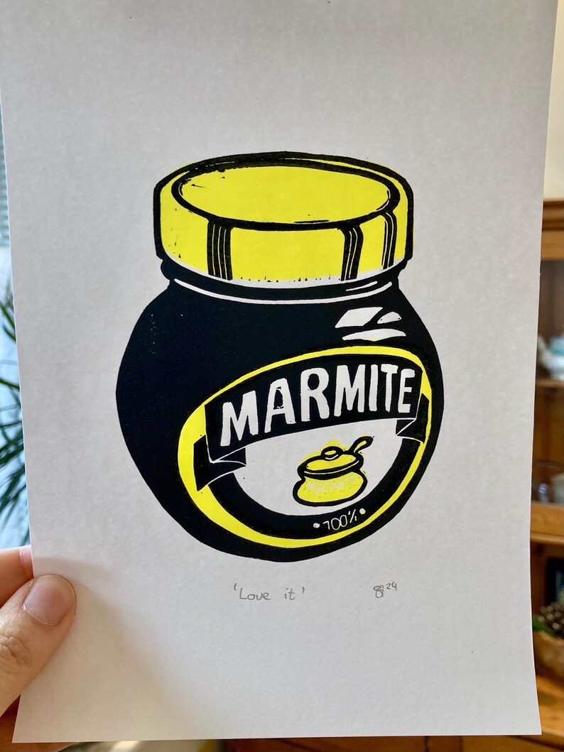 May include: A black and yellow linocut print of a Marmite jar. The jar has a yellow lid and the text "Marmite" in white letters. The text "100%" is printed in black below a yellow pot with a spoon. The text "'Love it'" is printed in black at the bottom of the print.