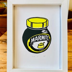May include: A framed print of a black and yellow Marmite jar with the text "Love it! You?" printed below. The jar has a yellow lid and a yellow label with the word "Marmite" in black letters. The label also has a small yellow pot with a spoon inside and the text "100%".