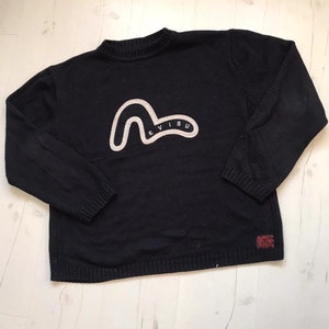 May include: A black knitted jumper with a white Evisu logo on the front. The logo is a stylised wave with the word "Evisu" written inside.