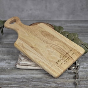 Op de afbeelding: Een houten snijplank met een handvat. De plank is gegraveerd met de tekst "your names" en een kleine huisillustratie. De tekst "Home Sweet Home" en de datum "21/11/2019" zijn ook gegraveerd op de plank.