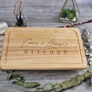 Könnte beinhalten: Rechteckiges Schneidebrett aus hellem Holz mit Saftrille. Die Worte "Corrie & George's KITCHEN" sind eingraviert. Das Brett liegt auf einer grauen Holzoberfläche.