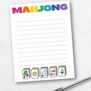 May include: White notepad with the word "MAHJONG" in rainbow-colored letters. The notepad has horizontal lines and illustrations of Mahjong tiles at the bottom. A pen rests on the surface.