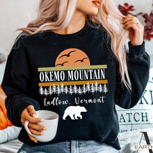 May include: Black sweatshirt with a graphic design featuring a sunset, trees, and a bear. The text "Okemo Mountain Ludlow, Vermont" is printed on the sweatshirt.