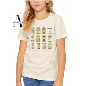 Pode incluir: Camiseta creme com uma grade de potes de picles ilustrados. Os potes variam em forma e design de r&oacute;tulo, mostrando uma variedade de tipos de picles. A palavra "EARTH" &eacute; impressa no lado esquerdo da camiseta.