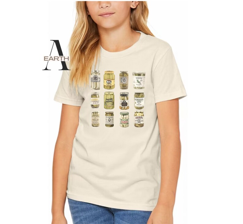 Pode incluir: Camiseta creme com uma grade de potes de picles ilustrados. Os potes variam em forma e design de r&oacute;tulo, mostrando uma variedade de tipos de picles. A palavra "EARTH" &eacute; impressa no lado esquerdo da camiseta.