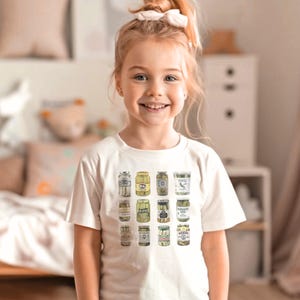 Pode incluir: Uma crian&ccedil;a vestindo uma camiseta creme com uma ilustra&ccedil;&atilde;o colorida de potes de picles. Os potes s&atilde;o dispostos em uma grade, mostrando diferentes tipos de picles. A crian&ccedil;a sorri para a c&acirc;mera, com um la&ccedil;o branco no cabelo.