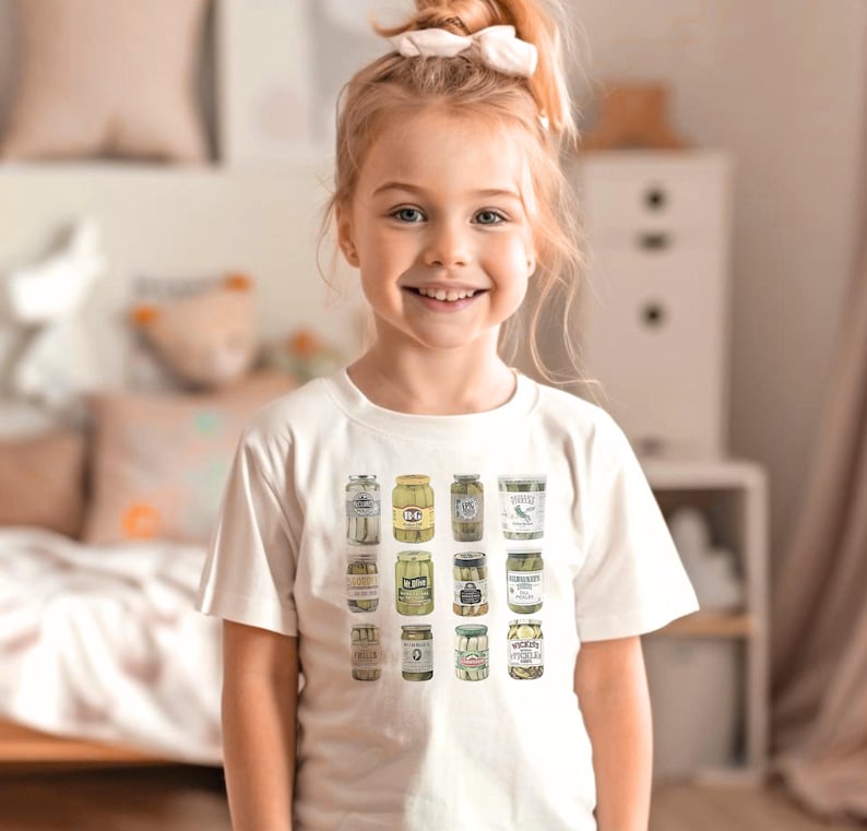 Pode incluir: Uma crian&ccedil;a vestindo uma camiseta creme com uma ilustra&ccedil;&atilde;o colorida de potes de picles. Os potes s&atilde;o dispostos em uma grade, mostrando diferentes tipos de picles. A crian&ccedil;a sorri para a c&acirc;mera, com um la&ccedil;o branco no cabelo.