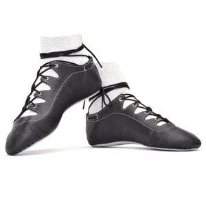 Puede incluir: Un par de zapatos de baile de cuero negro con costuras y cordones blancos. Los zapatos se usan con calcetines blancos y están diseñados para bailar. Sobre un fondo blanco.
