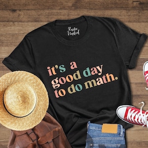 May include: Black t-shirt with the text "it's a good day to do math." in rainbow colors. The shirt is laid out on a wooden surface with a straw hat, a brown leather backpack, blue jeans, and red sneakers.