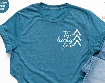 May include: A teal blue t-shirt with the text "The lucky few" and three white arrows on the front. The shirt is laid out on a white surface with a pair of white sneakers and a pair of light blue jeans.
