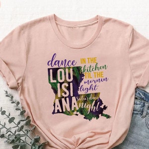 May include: A pink t-shirt with a graphic of the state of Louisiana. The graphic includes the text "dance in the kitchen til the mornin light Saturday night" and the words "LouisiAna".