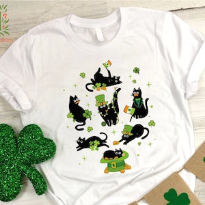 May include: White t-shirt with a black cat design. The cats are wearing shamrock hats and holding shamrocks. The shirt has a green shamrock design around the cats.
