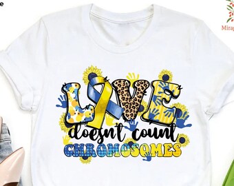 May include: White t-shirt with a graphic design of the word "LOVE" in blue, yellow, and black with sunflowers and handprints. The text "doesn't count chromosomes" is below the word "LOVE".