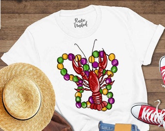 May include: White t-shirt with a red crawfish wearing purple, green, and gold Mardi Gras beads. The shirt is laid out on a wooden surface with a straw hat, a brown leather backpack, and red sneakers.