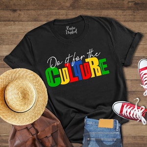 May include: Black t-shirt with the text "Do it for the CULTURE" in colorful letters. The letters are green, yellow, red, and blue. The shirt is laid out on a wooden surface with a straw hat, a brown leather backpack, and a pair of red sneakers.