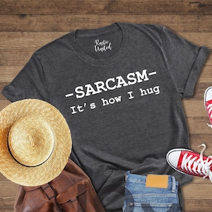 May include: A dark heather grey t-shirt with the text "SARCASM - It's how I hug" printed in white. The shirt is laid flat on a wooden surface with a straw hat, a brown leather backpack, a pair of red sneakers, and blue jeans.