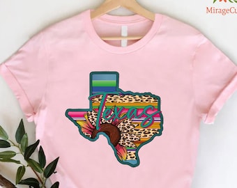 May include: A light pink t-shirt featuring a colorful graphic of the state of Texas. The graphic includes a sunflower, leopard print, and the word "Texas" in a turquoise script. The shirt is hanging on a wooden hanger.