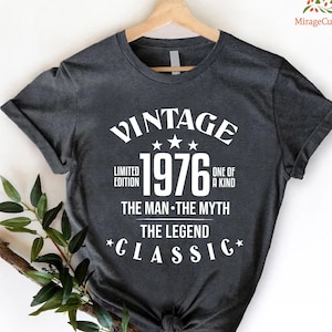 May include: Dark gray t-shirt with the text "VINTAGE 1976" in white, along with the phrases "LIMITED EDITION," "ONE OF A KIND," "THE MAN THE MYTH THE LEGEND," and "CLASSIC." The shirt is hanging on a wooden hanger.