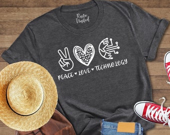 May include: A dark grey heather t-shirt with a white peace sign, heart, and the words "Peace Love Technology". The shirt is laid out on a wooden surface with a straw hat, a brown leather backpack, and a pair of red sneakers.