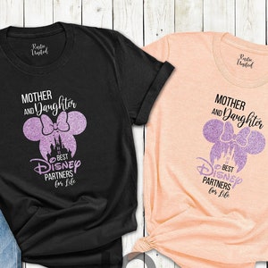 Puede incluir: Dos camisetas, una negra y otra melocot&oacute;n, con el texto "Mother and Daughter Best Disney Partners for Life". El dise&ntilde;o presenta una silueta de Minnie Mouse con un castillo. Tambi&eacute;n se ven unos vaqueros azules y gafas de sol.