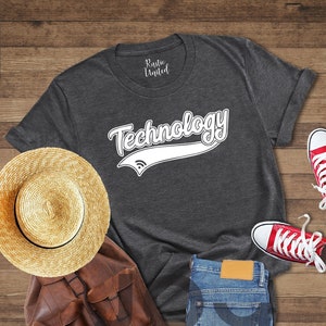 May include: A dark heather grey t-shirt with the word "Technology" in a white vintage-style font with a white baseball-style arc underneath. The shirt is laid out on a wooden surface with a straw hat, a brown leather backpack, blue jeans, and red sneakers.
