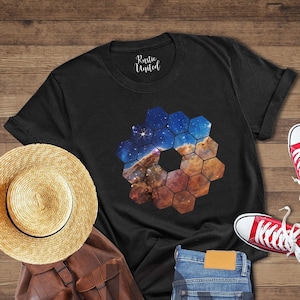 May include: Black t-shirt with a hexagonal design featuring a colorful nebula. The design is centered on the shirt. The shirt is laid out on a wooden surface with a straw hat, a brown leather backpack, a pair of red sneakers, and blue jeans.