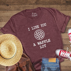 Puede incluir: Una camiseta de color marr&oacute;n jaspeado con el texto "I like you a waffle lot" y un gr&aacute;fico de waffle. La camiseta est&aacute; colocada sobre una superficie de madera con un sombrero de paja, una bolsa de cuero marr&oacute;n y zapatillas rojas.