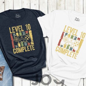 May include: Two t-shirts with a retro-style video game controller graphic and the text "Level 10 Complete". One shirt is navy blue and the other is white.