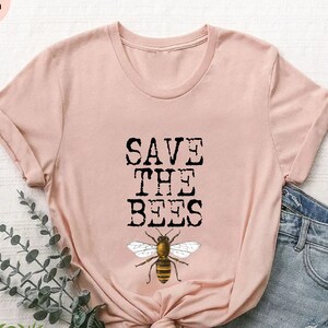 May include: A pink t-shirt with the text "SAVE THE BEES" in black lettering and a graphic of a bee. The shirt is on a white surface with a pair of blue denim shorts and green foliage.