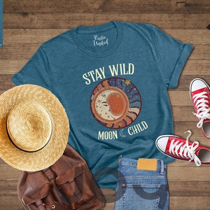 May include: A teal blue t-shirt with the text "Stay Wild Moon Child" and a graphic of a crescent moon with stars. The shirt is laid out on a wooden surface with a straw hat, a brown leather backpack, and red trainers.